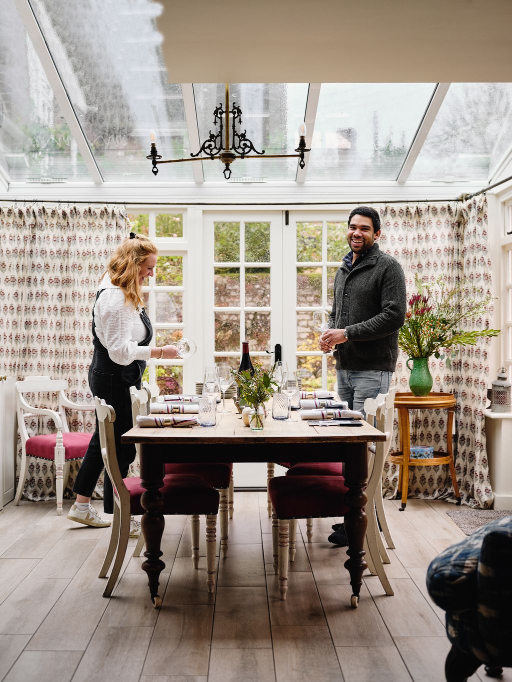 The couple set the table ready for a family gathering in front of curtains in Robert Kimes ‘Field Poppy on muslin.