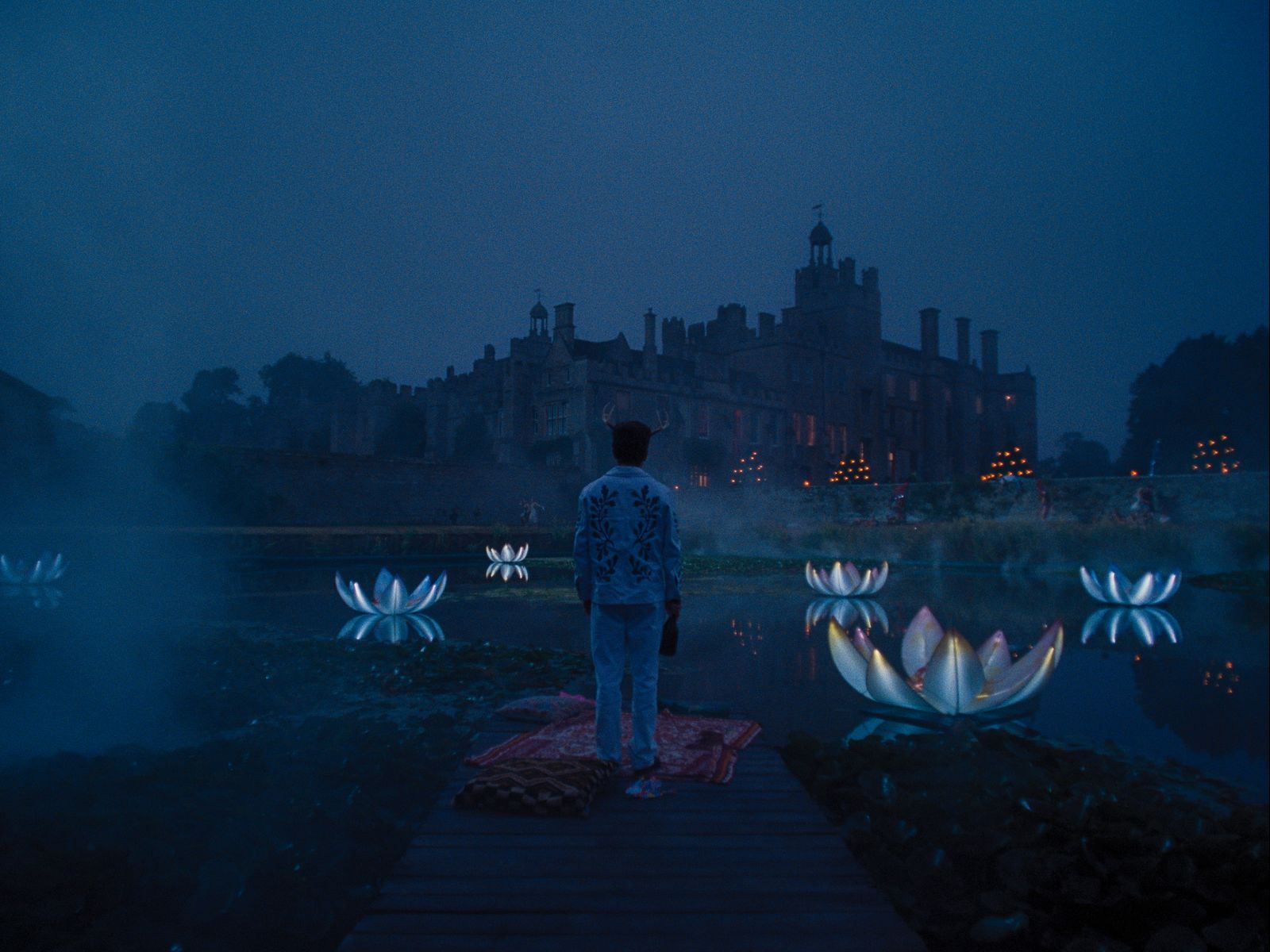 Dark still from Saltburn of mist over pond Barry Keoghan in foreground with back to camera estate in background lotus...