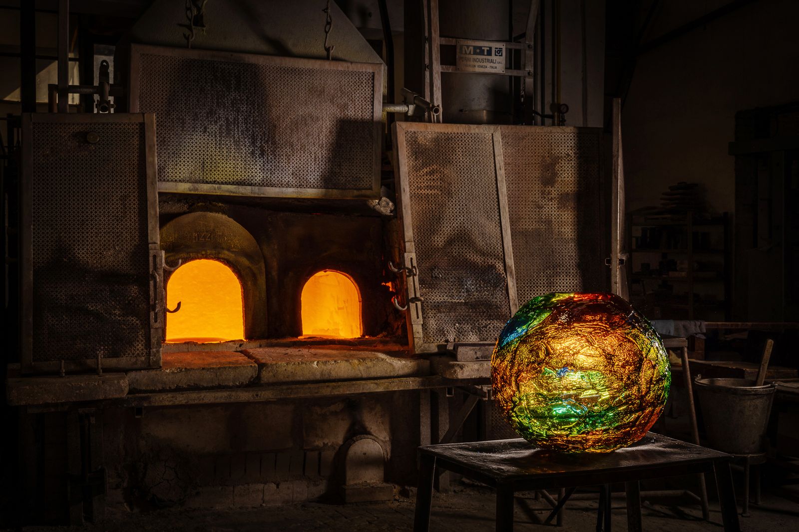 A glowing glass form cools beside the roaring furnaces.